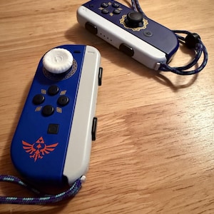 May include: Two blue and gray game controllers with a Legend of Zelda theme. The controllers have black buttons and a white joystick. One controller has a red and orange emblem. Both controllers have blue and purple wrist straps.