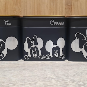 Può includere: Tre barattoli neri con coperchi in bambù, etichettati "Tea", "Coffee" e "Sugar". Ogni barattolo presenta un design argentato di Topolino o Minnie. I barattoli sono disposti su una superficie chiara.