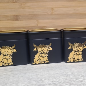 May include: Three navy blue metal storage tins with gold-coloured lids. Each tin features a gold-coloured illustration of a Highland cow. The tins are square-shaped and sit on a light-coloured surface.