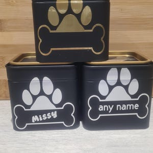 Può includere: Contenitori quadrati neri con bordi dorati e disegni di impronte di zampe. Uno ha un'impronta di zampa e un osso dorati, mentre gli altri hanno impronte di zampe e ossa bianche con i nomi "Missy" e "any name".