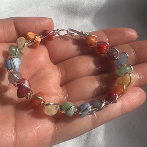 May include: A handmade bracelet featuring a variety of colorful gemstones, including red, orange, yellow, green, blue, and purple. The stones are wire-wrapped in a silver-colored metal, creating a unique and eye-catching design. The bracelet is a perfect accessory for any outfit.