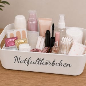 Könnte beinhalten: Ein weißer, rechteckiger Kunststoff-Aufbewahrungskorb mit integrierten Griffen und dem grauen Schriftzug Notfallkörbchen auf der Vorderseite. Der Korb ist mit verschiedenen Körperpflegeartikeln gefüllt, darunter Flaschen, Tuben, Wattestäbchen und Haaraccessoires.
