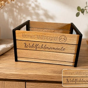 Könnte beinhalten: Eine rechteckige Holzkiste mit schwarzen Metallrahmen an den Seiten. Die Vorderseite zeigt den eingravierten Text "kleines Mitbringsel" neben einem lächelnden Gesichtssymbol sowie den Text "Wohlfühlmomente" in Schreibschrift darunter.
