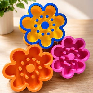 Könnte beinhalten: Drei blütenförmige Seifenschalen in Gelb, Orange und Pink. Die gelbe Schale hat blaue Akzente, während die orangefarbenen und pinkfarbenen Schalen passende Mittelpunkte haben. Jede Schale hat Stifte zur Entwässerung.