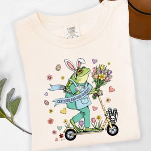 May include: Cream-colored t-shirt featuring a cartoon frog wearing bunny ears, scrubs, and riding a scooter while holding flowers. The frog has a banner that reads "CERTIFIED RAD-EGG-OLOGIST". Decorative flowers and hearts surround the frog.