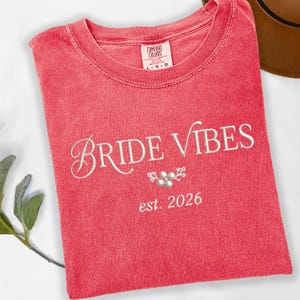 May include: A coral-colored t-shirt with the words "Bride Vibes" embroidered in a cursive font. Below the text is a small floral design and the date "est. 2026". The shirt has a crew neck and short sleeves.