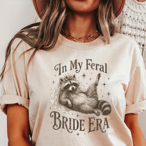 May include: Beige t-shirt featuring a raccoon wearing a veil and crown, with the text "In My Feral Bride Era". The raccoon is lying down with one paw raised. The design includes star accents.