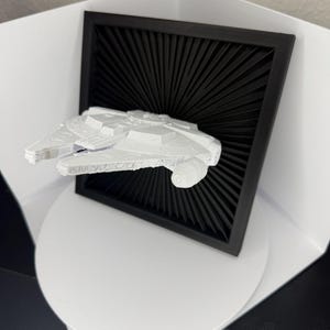 Könnte beinhalten: Ein weißes Modell des Raumschiffs Millennium Falcon, vor einem schwarzen, strahlenförmigen Hintergrund in einem schwarzen Rahmen. Das Modell befindet sich auf einer weißen, kreisförmigen Plattform, wodurch ein 3D-Effekt entsteht. Das Thema ist Science-Fiction.