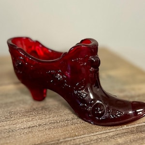 Puede incluir: Una figura de zapato de vidrio rojo intenso que presenta un patrón floral moldeado con rosas y hojas en relieve en la superficie exterior. El objeto tiene una punta afilada, un tacón pequeño y una abertura festoneada, hecho de vidrio rubí translúcido.