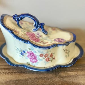 May include: A cream-colored ceramic butter dish with a scalloped edge and a blue rim. The lid features a decorative blue handle and a floral pattern with pink, red, and blue flowers. The base is also cream-colored with a matching blue scalloped rim.