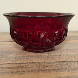 May include: A deep red glass bowl featuring a prominent geometric diamond pattern across its exterior surface. The bowl has a wide, flared rim and a circular base, with the translucent red glass catching the light to highlight the faceted texture.