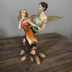 Puede incluir: Una figura de dos hadas con alas translúcidas sobre una base de tocón de árbol. Un hada viste un vestido naranja y la otra una túnica verde. La base es marrón con pequeñas flores rosas adheridas al costado del tronco del árbol.