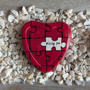 Könnte beinhalten: Ein rotes herzförmiges Puzzleteil mit den Worten "Missing you..." auf einem weißen Teil. Das Puzzle ist von kleinen, weißen Steinen umgeben. Das Herz hat schwarze Linien, um die Puzzleteile zu definieren.