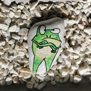 May include: A hand-painted, white rock featuring a green frog illustration with black outlines. The frog has large eyes and is drawn with a cartoonish style. The rock is surrounded by small, white pebbles.