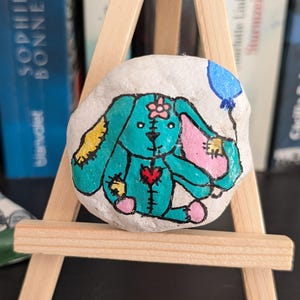 May include: A hand-painted rock with a teal bunny design, pink and yellow details, and a blue balloon. The bunny has a heart and flower details. The rock is displayed on a small wooden easel, a charming decorative item.