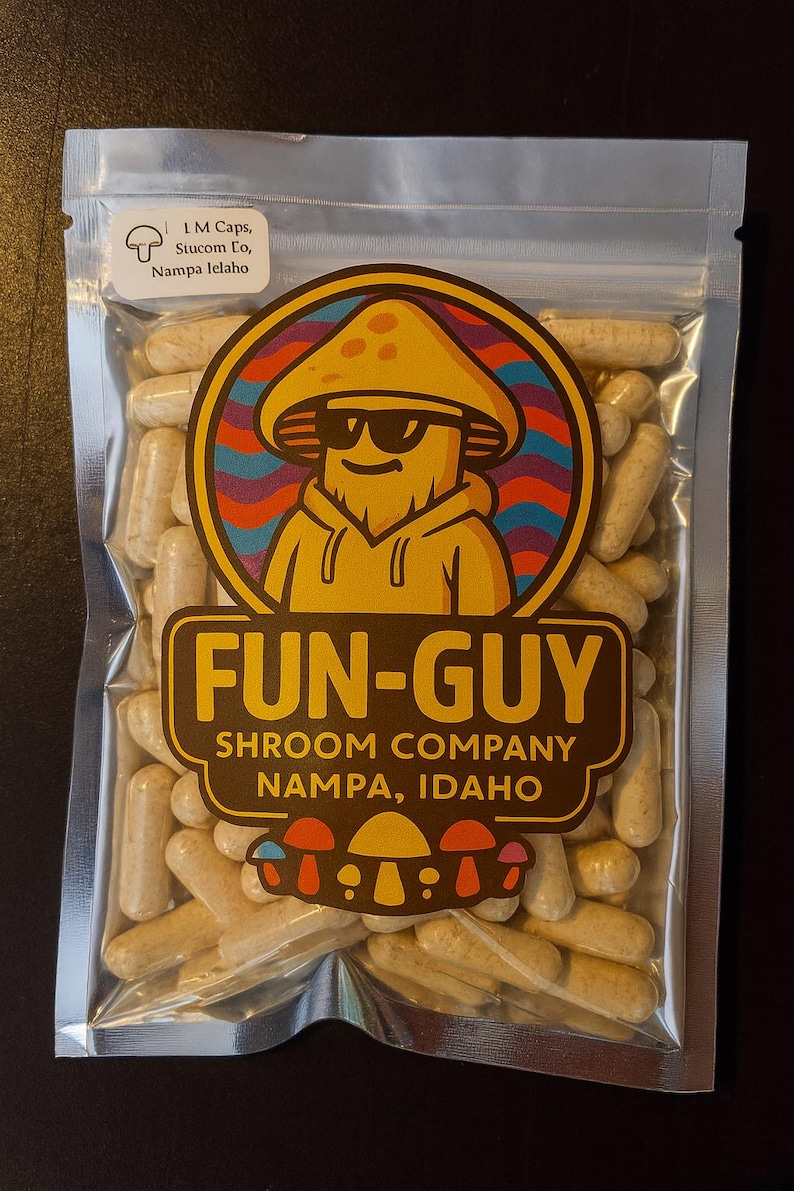 Op de afbeelding: Een transparante plastic zak gevuld met beige capsules. De zak heeft een kleurrijk logo met een paddenstoelpersonage met een zonnebril en de tekst "FUN-GUY SHROOM COMPANY NAMPA, IDAHO".