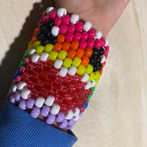 Może przedstawiać: Kolorowa bransoletka kandi z koralikami w odcieniach różu, pomarańczu, żółci, zieleni, czerni, bieli i fioletu. Bransoletka jest noszona na nadgarstku, a koraliki ułożone są we wzór.