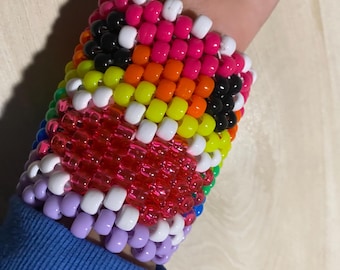 Può includere: Un braccialetto kandi colorato con perline rosa, arancioni, gialle, verdi, nere, bianche e viola. Il braccialetto è indossato su un polso, con le perline disposte in un motivo.