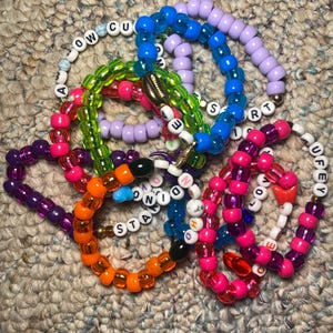 May include: A collection of colorful beaded bracelets in various shades of blue, purple, green, orange, and pink. Some bracelets feature white letter beads spelling out words like "LOVE" and "STAY". The bracelets are made of small, round beads.