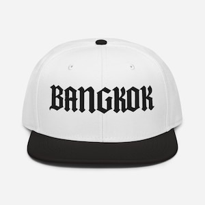 Può includere: Un cappellino snapback bianco con visiera nera e un bottone nero sulla parte superiore. La parte anteriore del cappellino presenta la parola BANGKOK ricamata in un carattere gotico nero e audace al centro della corona bianca.
