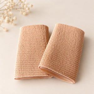 May include: Two beige finger sleeves made of a flexible, knit fabric. Each sleeve features a textured, woven exterior surface and a smooth, semi-transparent gel-like lining visible at the open end, designed for finger protection.