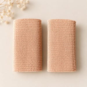 May include: Two rectangular, light peach-colored wristbands featuring a dense, uniform knit texture. The bands are positioned side-by-side, displaying a consistent, flexible fabric construction with a subtle, fine-grained surface pattern throughout.