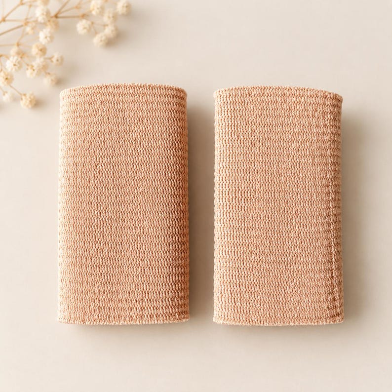 May include: Two rectangular, light peach-colored wristbands featuring a dense, uniform knit texture. The bands are positioned side-by-side, displaying a consistent, flexible fabric construction with a subtle, fine-grained surface pattern throughout.