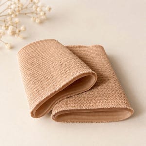 May include: A pair of beige compression sleeves featuring a textured, knit-like exterior surface and a smooth, solid-colored interior lining. The sleeves are cylindrical in shape and have a consistent, neutral tan color throughout the fabric.