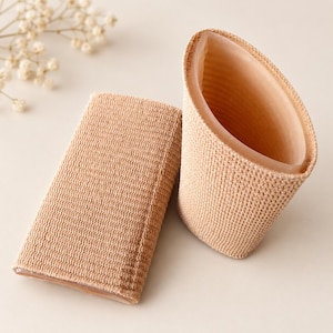 May include: Two beige, textured, cylindrical fabric sleeves with a soft, smooth interior lining. One sleeve is flattened into a rectangular shape, while the other remains open in a tubular form, showcasing the contrast between the exterior knit.