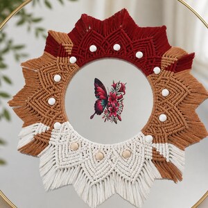 May include: A circular mirror featuring a macrame frame in red, brown, and white. The frame has a starburst design with white and beige details. A butterfly and floral design is reflected in the mirror's centre.