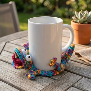 May include: A plain white ceramic mug sits inside a colorful, hand-crocheted lizard-shaped coaster. The lizard features a vibrant mix of blue, pink, yellow, and purple yarn, with two white protruding eyes and an open mouth showing a red tongue.