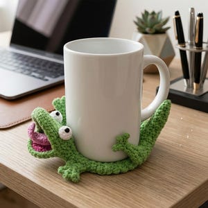 May include: A white ceramic mug resting inside a green crocheted coaster shaped like a lizard. The lizard figure features a wide-open mouth with a pink tongue, two white eyes with black pupils, and four small limbs that wrap around the mug base.