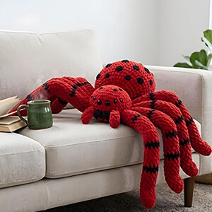 Puede incluir: Un peluche de araña grande, rojo y negro, con una araña más pequeña. Las arañas tienen manchas y rayas negras. Los peluches están sobre un sofá blanco. También se ve una taza verde y un libro.