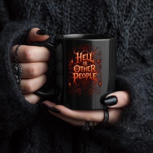 Può includere: Tazza in ceramica nera con la scritta "Hell is Other People" in un carattere gotico rosso fuoco. La tazza ha un manico nero ed è tenuta da una persona che indossa un maglione nero e anelli.