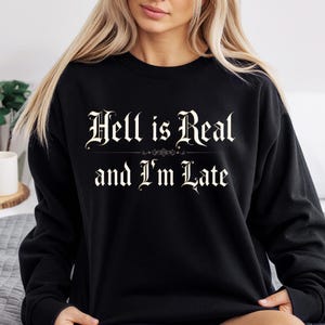 以下が含まれることがあります： 黒色のスウェットシャツ。白いゴシック体で「Hell is Real and I'm Late」と書かれています。テキストは中央に配置されています。クルーネックのデザインで、カジュアルなスタイルです。
