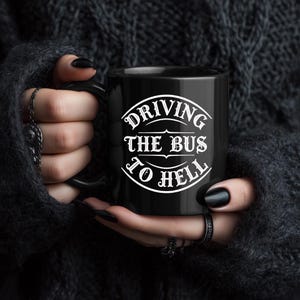 Può includere: Tazza in ceramica nera con la scritta bianca "DRIVING THE BUS TO HELL", tenuta da una persona. La tazza ha un manico nero e una finitura lucida. Il testo è in un carattere stilizzato.