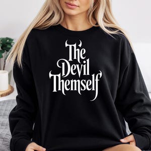 May include: A white ceramic mug with a black handle and the text "But First, Coffee" in a bold, black font. The mug is set against a blurred background, suggesting a cosy setting.