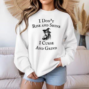 May include: White sweatshirt with the text "I Don't Rise and Shine, I Curse and Grind" in black. The graphic features a witch with a coffee cup. The sweatshirt is a casual, comfortable style.