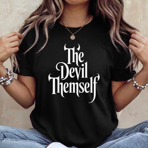 Può includere: T-shirt nero con la scritta bianca "The Devil Themself" in un font gotico. La maglietta ha maniche corte e girocollo. La modella indossa jeans blu.
