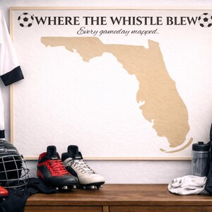 Może przedstawiać: Oprawiona mapa Florydy z napisem "WHERE THE WHISTLE BLEW" i "Every gameday mapped". Piłka nożna, koszulka, korki i kask leżą na drewnianej powierzchni. Obecna jest również czarna torba sportowa i butelka wody.
