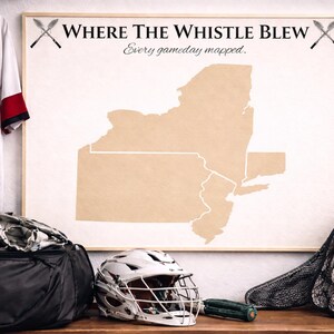Puede incluir: Un mapa enmarcado del estado de Nueva York con el texto "Where The Whistle Blew" y "Every gameday mapped." También se ven una camiseta de lacrosse, un casco, guantes y pelotas. La camiseta tiene el número 22.