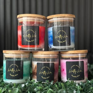 May include: Five glass jar candles with wooden lids are arranged on a bed of green foliage. Each candle has a different color wax, including red, blue, green, brown, and pink. The candles have black labels with gold text that reads "Luxury Scents Wealthy Wax Paraffin Wax".
