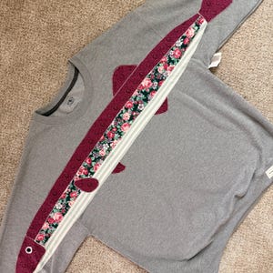 May include: A gray sweatshirt with a large fish applique. The fish is made of burgundy fabric with a floral pattern and white stripes. The fish has burgundy fins and a burgundy eye.