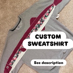 May include: A grey crewneck sweatshirt featuring a large fish-shaped appliqué across the front. The fish body is made of a dark maroon fabric with a floral pattern insert and a white eye. Text on the image reads CUSTOM SWEATSHIRT and See description.