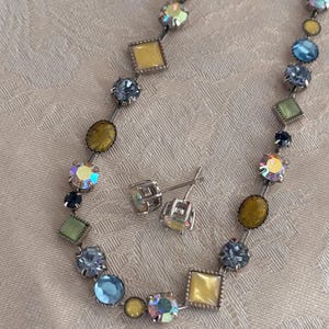 Op de afbeelding: Een ketting en bijpassende oorstekers set. De ketting heeft een verscheidenheid aan kleurrijke, gefacetteerde edelstenen in tinten blauw, geel, groen en iriserend. De stenen zijn gezet in een zilverkleurig metaal. De oorbellen hebben een vergelijkbaar ontwerp.