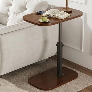May include: A dark brown, adjustable side table with a rectangular base and a curved top. A cup of coffee, a book, and a smartphone rest on the table. The table is next to a white sofa.