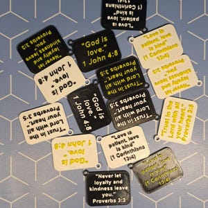 Könnte beinhalten: Eine Sammlung kleiner, quadratischer Schlüsselanhänger mit abgerundeten Ecken, die jeweils einen anderen Bibelvers in schwarzem oder gelbem Text auf weißem oder schwarzem Hintergrund tragen. Die Schlüsselanhänger haben ein kleines Loch zur Befestigung.
