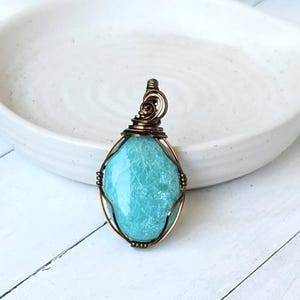 May include: A handcrafted pendant with a light blue gemstone, encased in copper wire. The stone has a textured surface and an oval shape. The copper wire is intricately woven around the stone, creating a unique piece of jewellery.