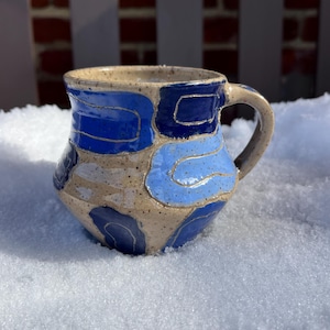 May include: A handmade ceramic mug with a beige base and blue abstract designs. The mug features a rounded body, a curved handle, and is approximately 4 inches tall. The blue patterns vary in shade and shape, adding a unique touch to this functional art piece.