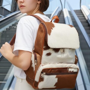May include: A brown and white cowhide backpack with a flap closure and a front zippered pocket. The backpack has brown leather straps and trim. The background shows an escalator and a building.